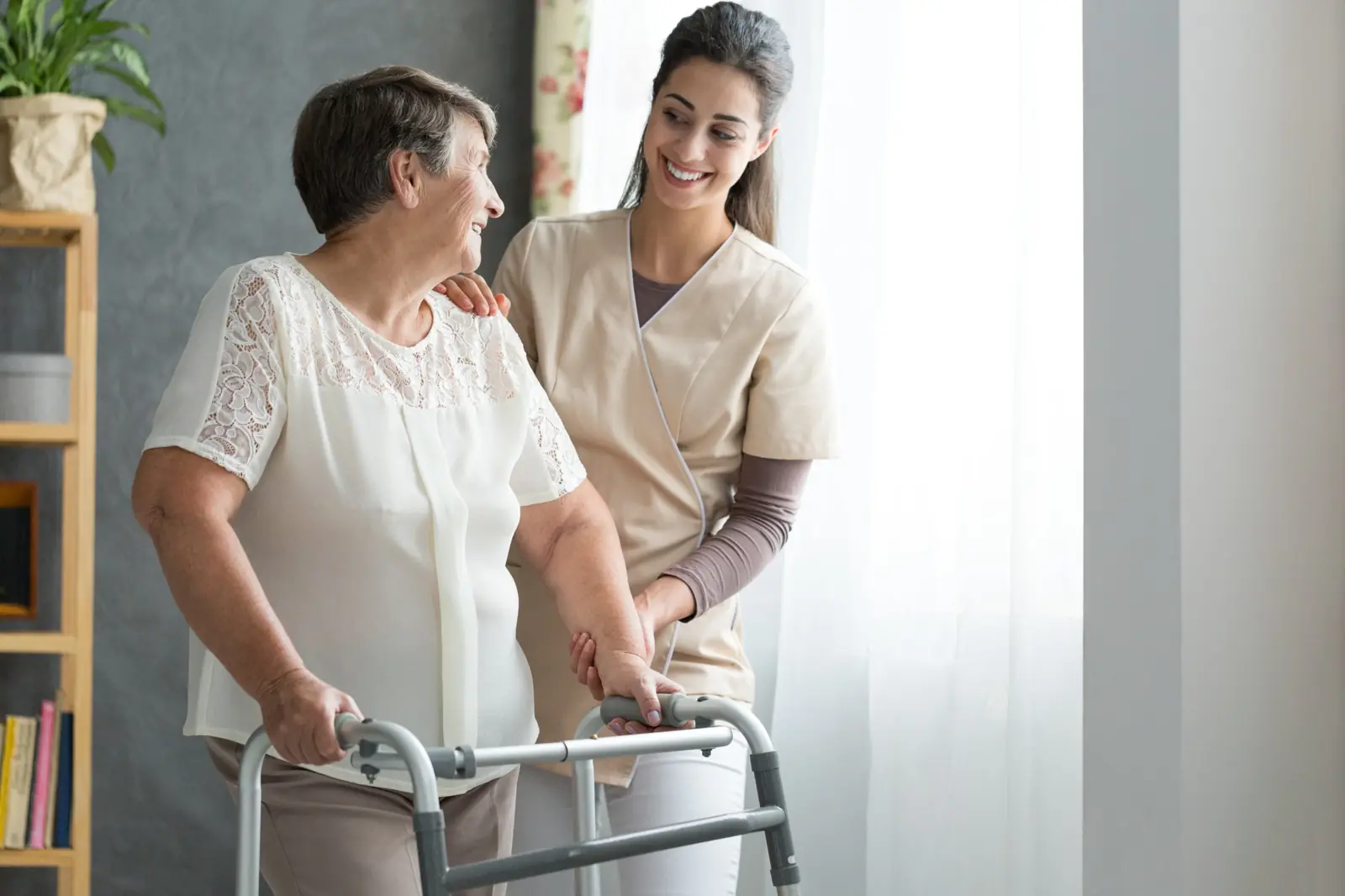 Home health nurse helping a smiling senior woman walk with a walker at home